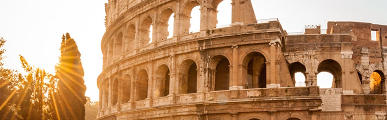 The sun shining next to the Coliseum, a round stone ruin with many arches and layers. 